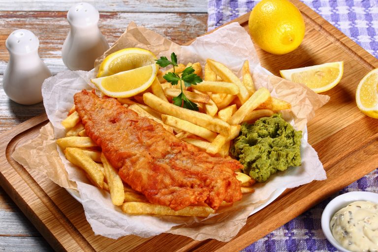 delicious crispy fish and chips - fried cod, french fries, lemon slices, tartar sauce and mushy peas on plate on paper. parsley and lemon wedges on wooden table, view from above, close-up